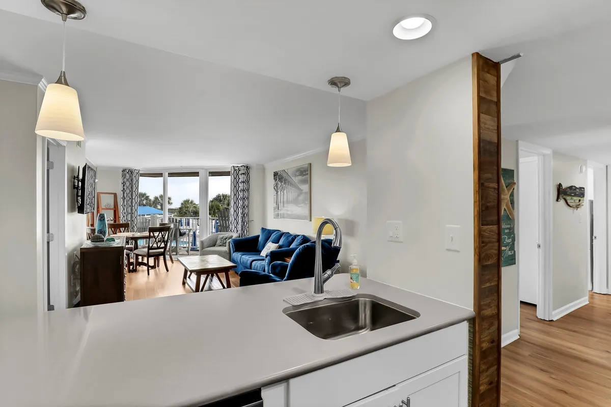 A bright, open-concept space connects the kitchen sink, a blue couch, dining table, and large windows, blending living, dining, and kitchen areas seamlessly. Coastal hues and sunlight enhance the welcoming atmosphere.