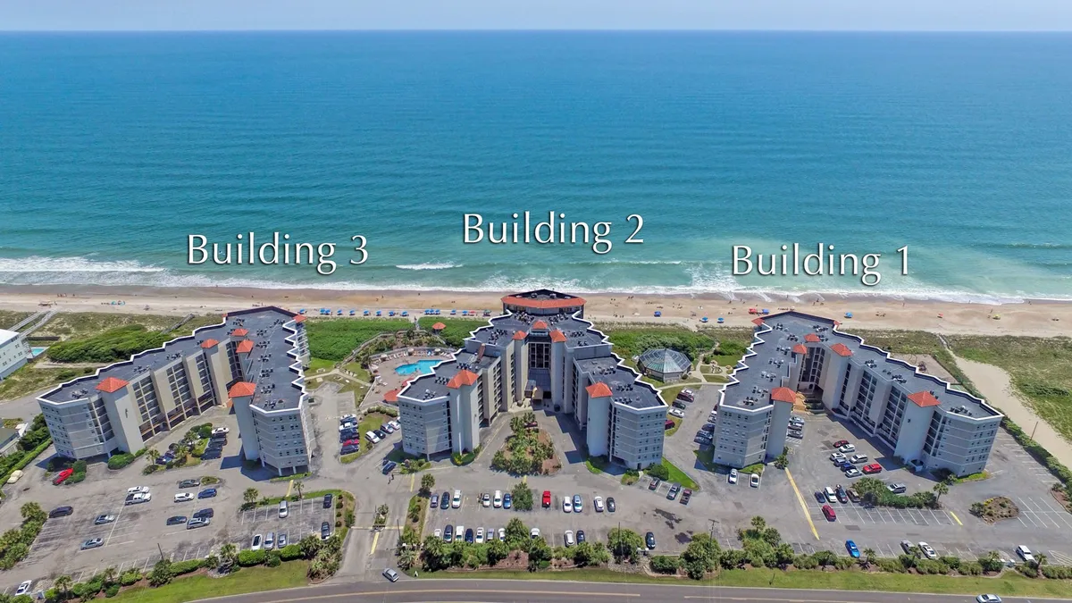 A broad exterior shot showing three resort buildings, a large swimming pool, and the ocean view beyond. Loungers and poolside areas are visible, emphasizing the blend of upscale accommodations and premier resort amenities, all with a stunning coastal backdrop.