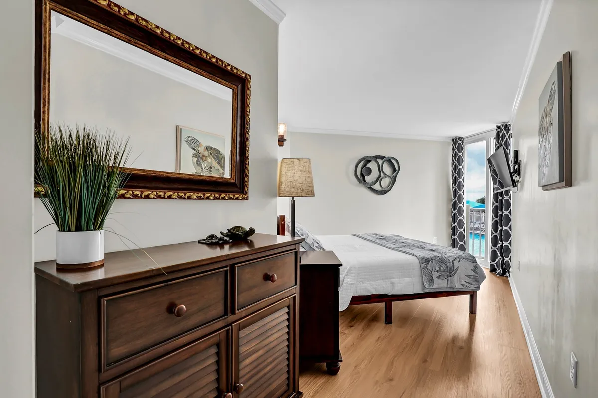 A tranquil bedroom featuring a bed with patterned bedding, a wooden dresser, mirror, and lamp. The room is softly lit, with wall decor adding a touch of personality. The presence of a TV suggests amenities for entertainment and relaxation.