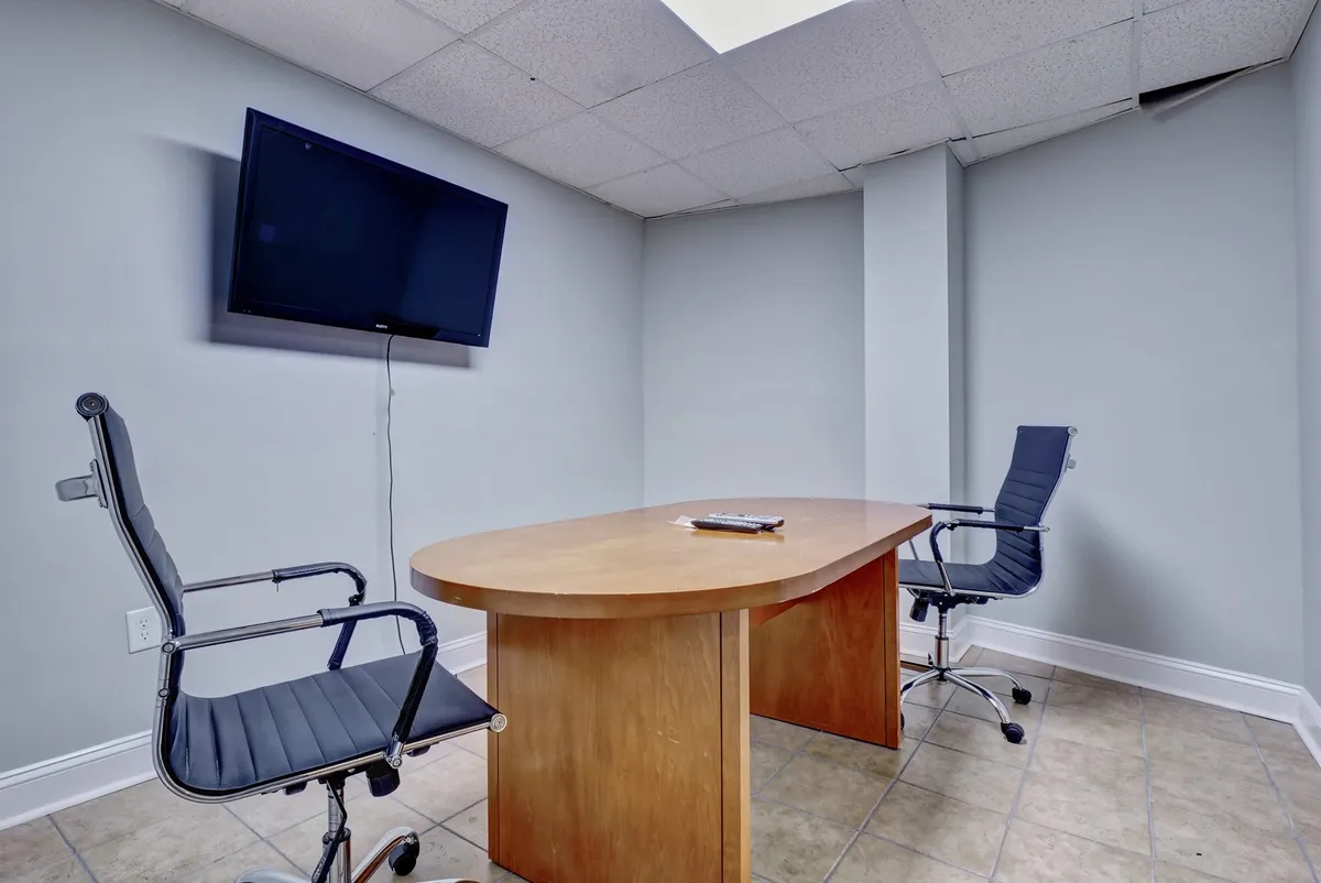 A professional business center setting, furnished with a large wooden conference table, two office chairs, and a wall-mounted television—ideal for meetings, video calls, or focused work sessions.