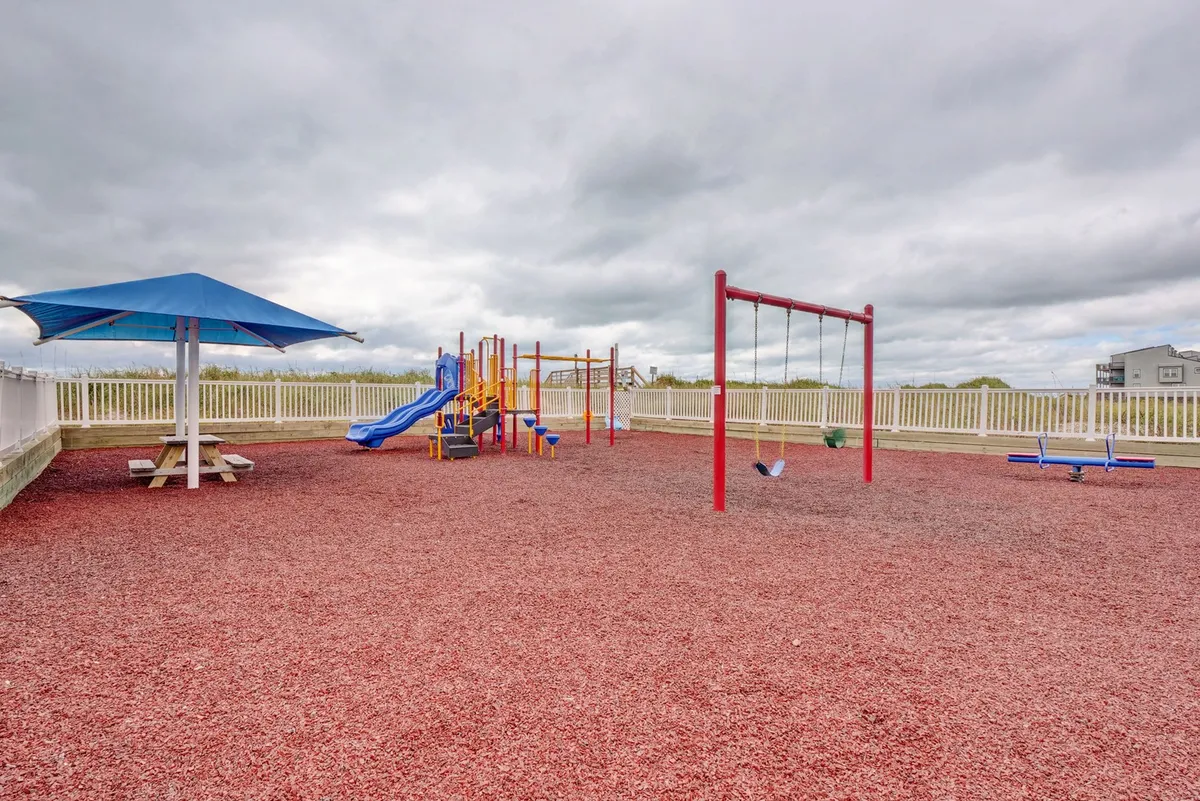 A vibrant outdoor playground scene with swings, climbing equipment under a canopy, and a picnic table in the background. Children are seen playing, while family and friends gather nearby, creating an atmosphere of joy and togetherness.