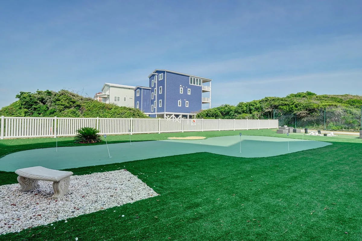 This image captures the cheerful putting green area with neat artificial turf, decorative white stone, a blue house in the background, and a white fence. A bench provides a spot for relaxation, and the area looks perfect for leisure activities under the sun.