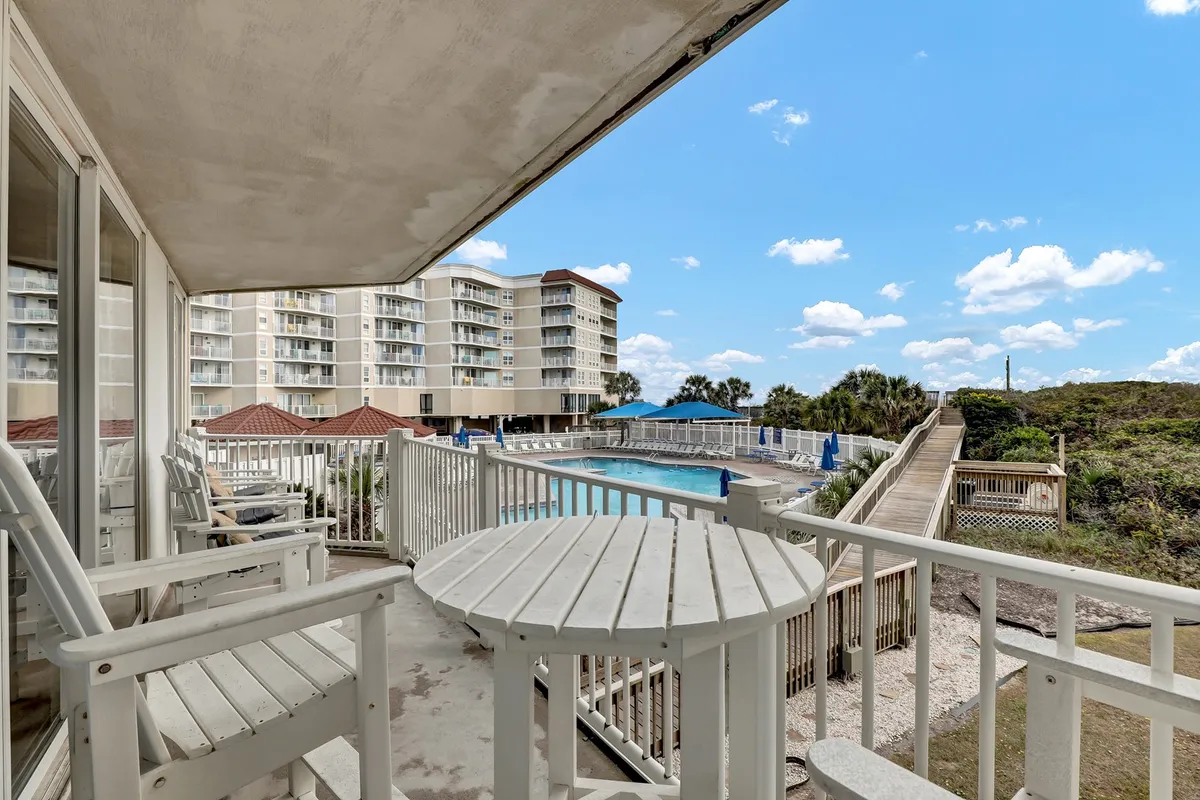 A wide exterior view showing the resort's sparkling swimming pool, surrounded by lush greenery and comfortable balcony seating. The hotel building stands in the background, and the pool area is inviting for both relaxation and social gatherings.
