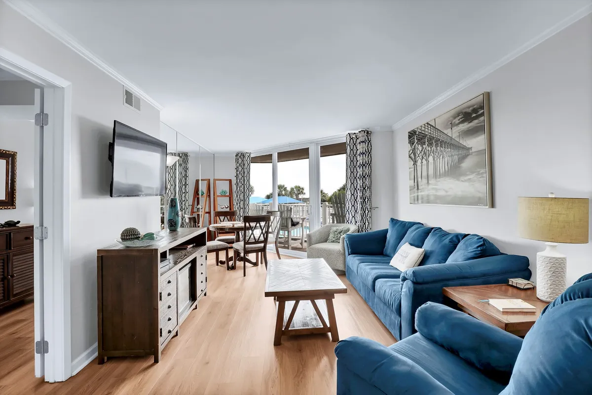 This image features a blue couch arranged around a wooden coffee table, with a smart TV mounted on the wall and large windows that allow natural light to fill the room. Decorative artwork and modern furnishings create a comfortable, contemporary ambiance.