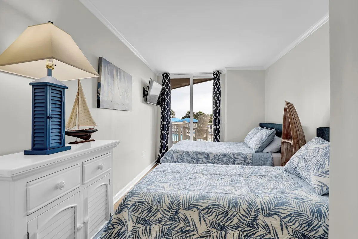 A bright bedroom with two beds, each dressed in blue leaf-patterned bedding. A white dresser with nautical decor and a lamp provide functional charm. The large sliding glass door opens to a balcony, inviting in natural light and offering an outdoor connection.