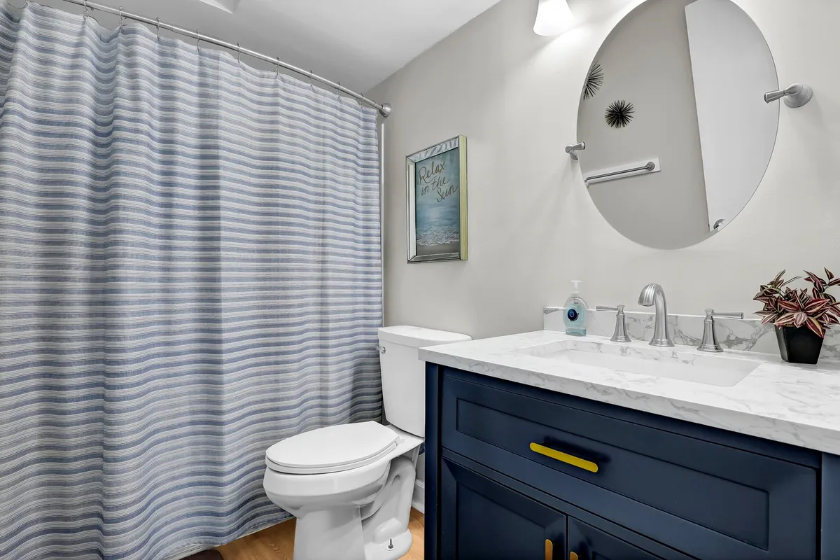 A chic, contemporary bathroom with a blue vanity, marble countertop, round mirror, and a striped shower curtain. The space is modern, fresh, and inviting, suggesting a pleasant setting for daily routines or unwinding after a long day.